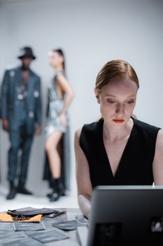 Caucasian female designer focuses intensely on laptop in stylish studio setting with models.