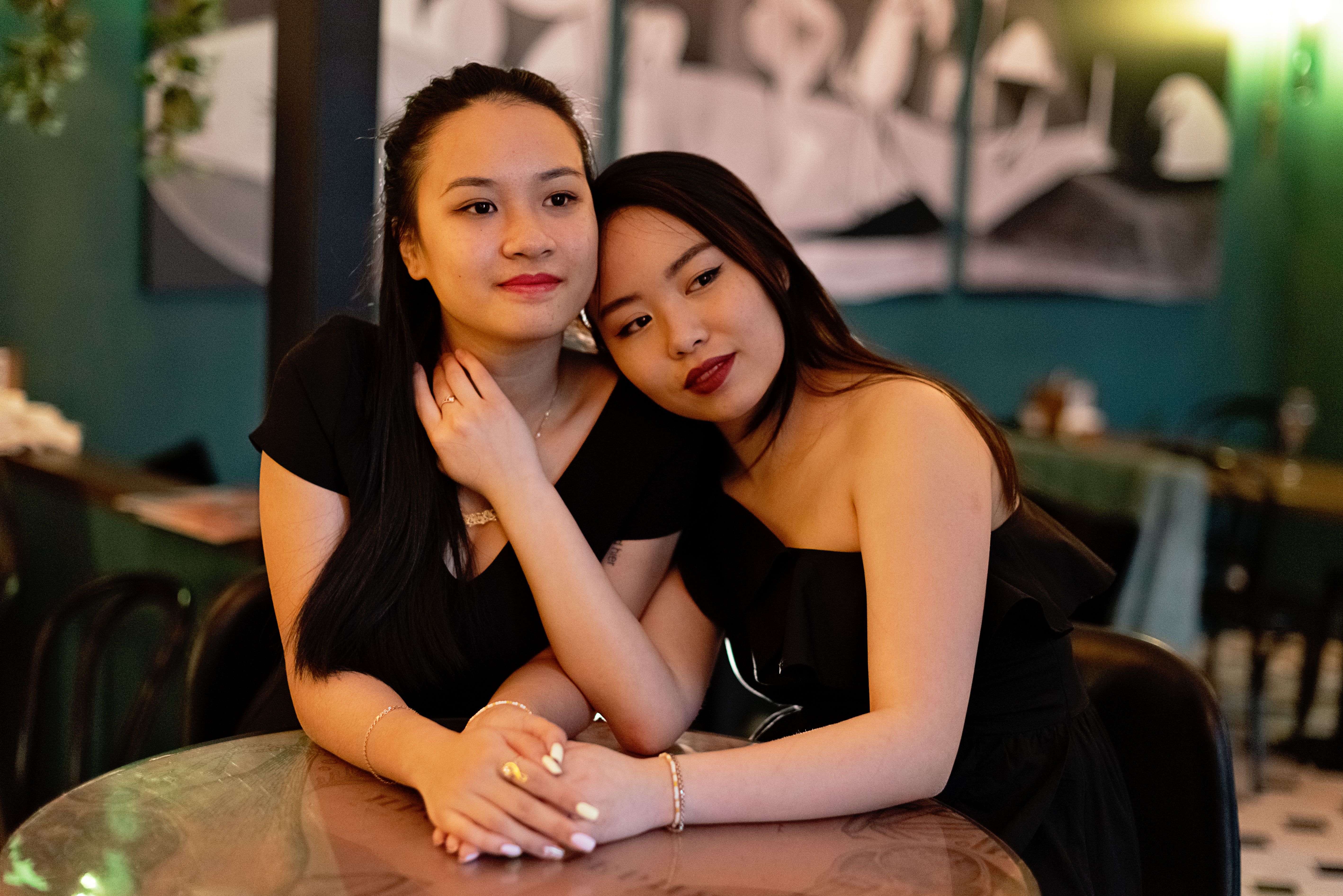 Two women in black dresses share a touching moment at a cafe, symbolizing love and companionship.