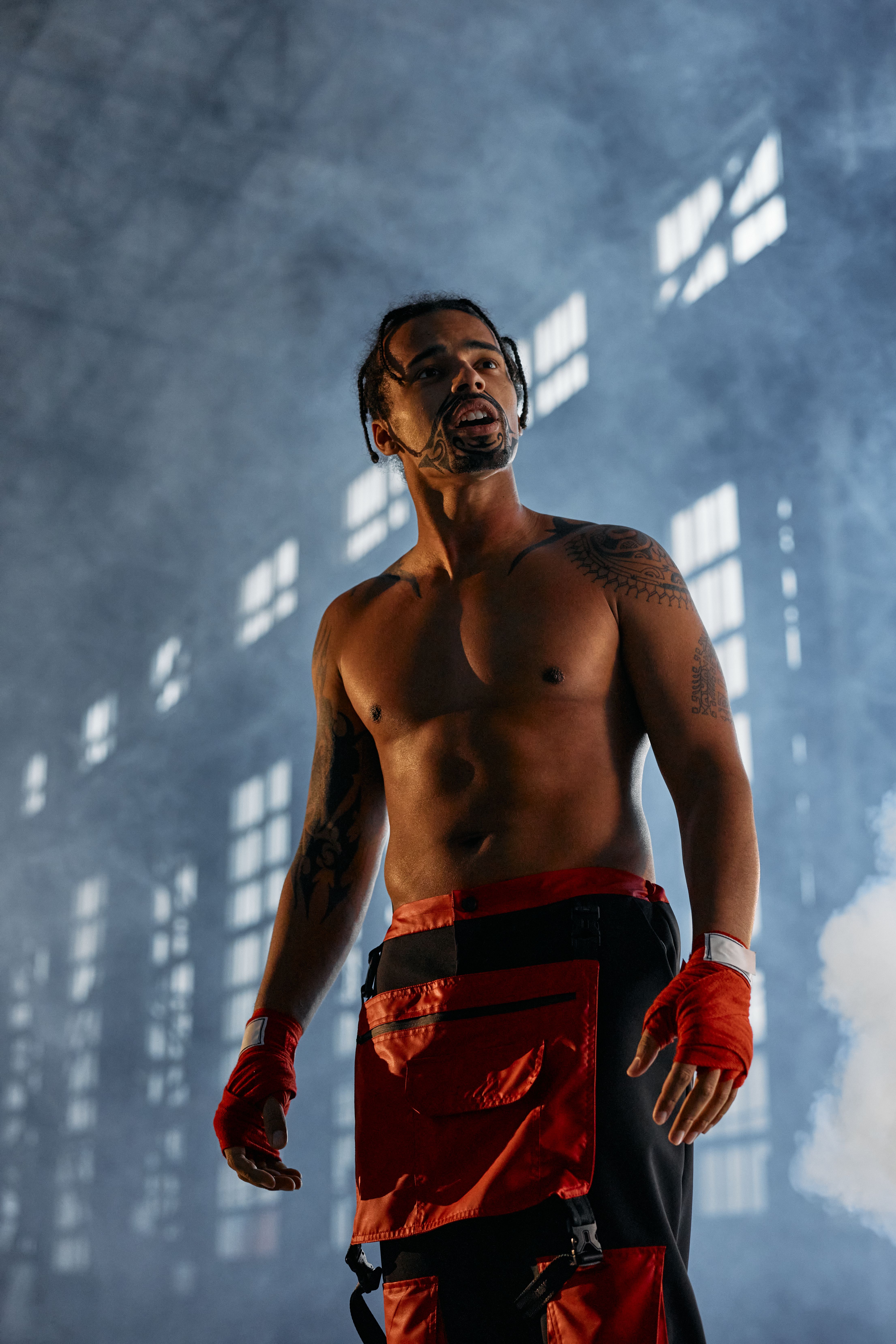 A shirtless, tattooed martial artist stands confidently in a smoky, dramatic indoor setting.