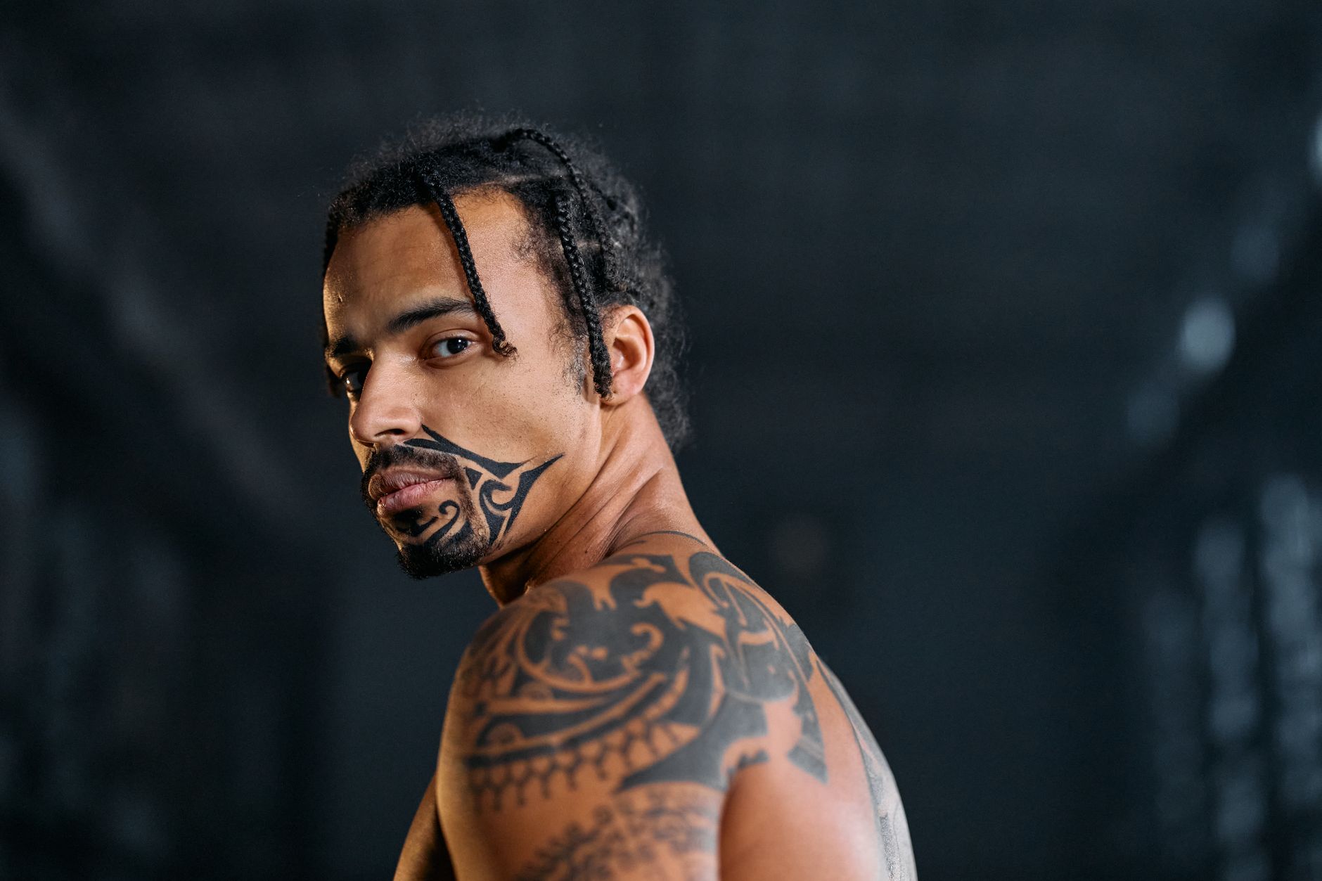 Portrait of a tattooed man with a face tattoo and dreadlocks in a dimly lit setting.