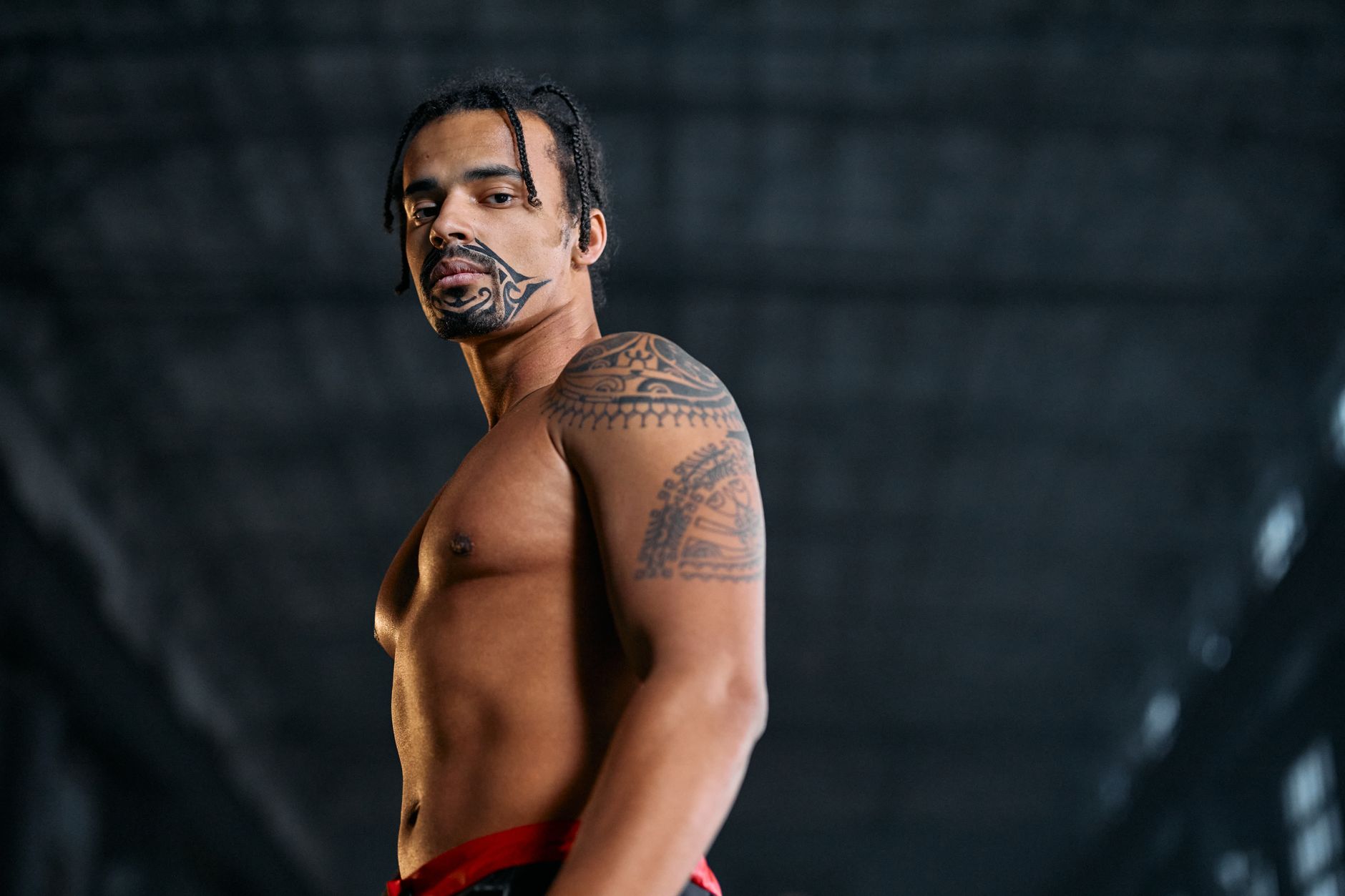 Stylish shirtless man with tattoos poses confidently in an industrial setting. Low-angle shot captures his cool demeanor.