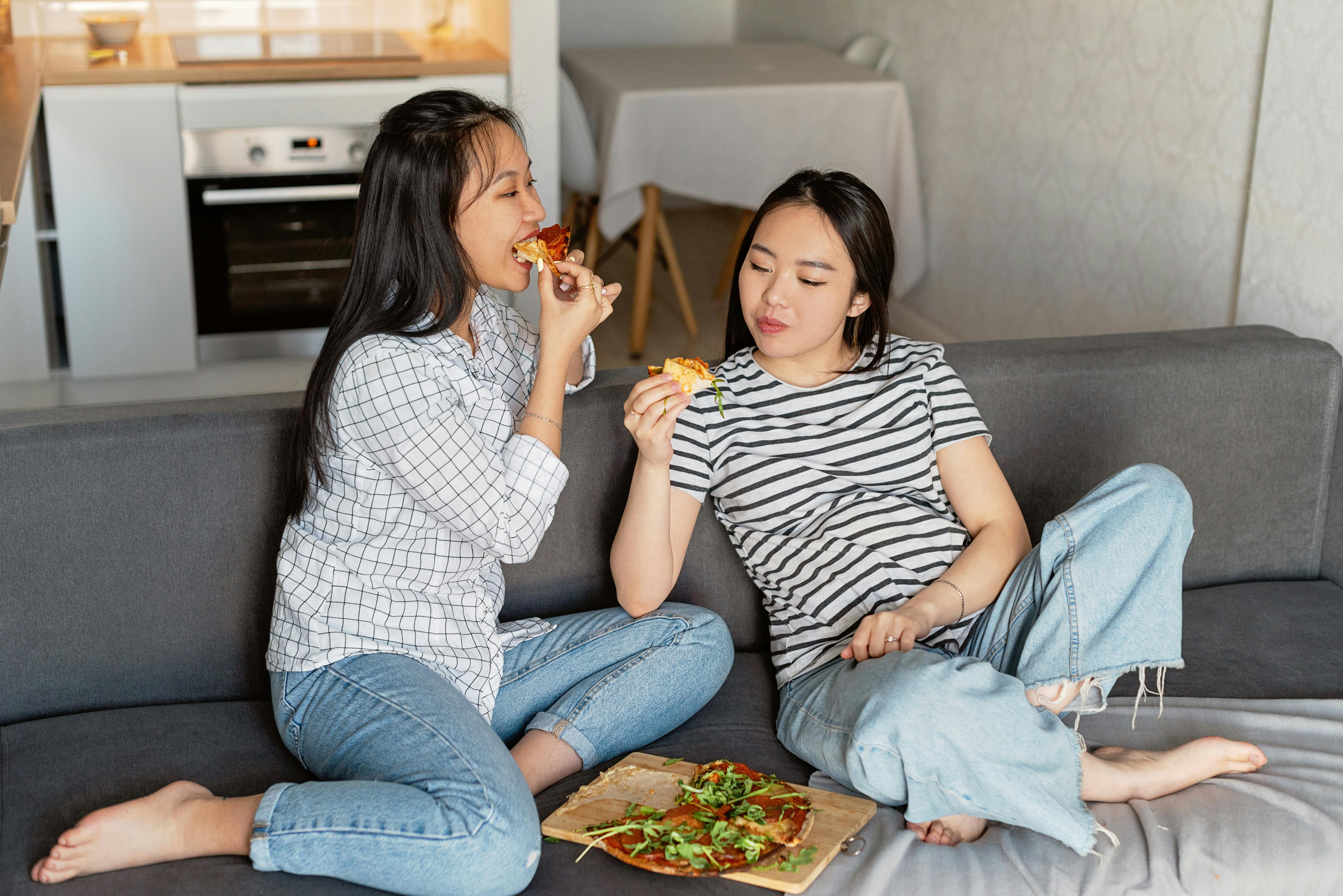 Two women sitting on a sofa at home, enjoying slices of pizza, embracing leisure time.