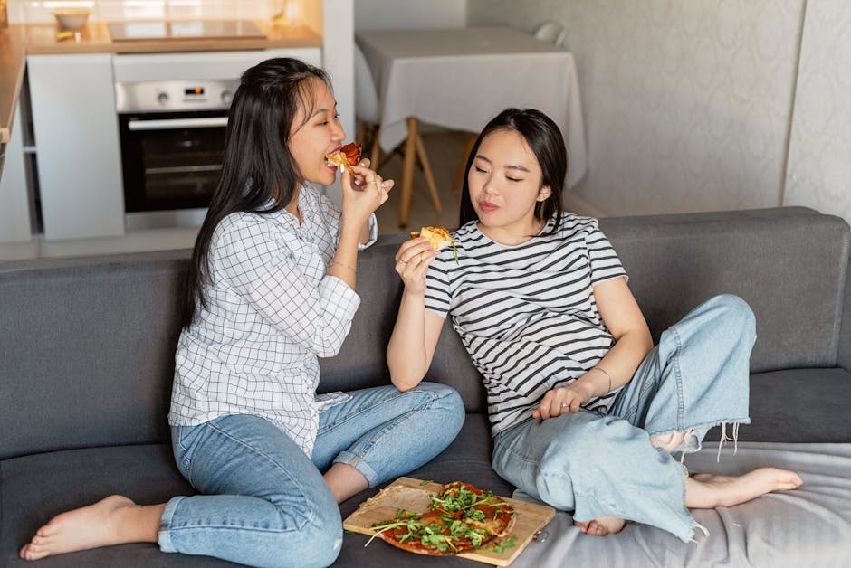 Two women sitting on a sofa at home, enjoying slices of pizza, embracing leisure time.