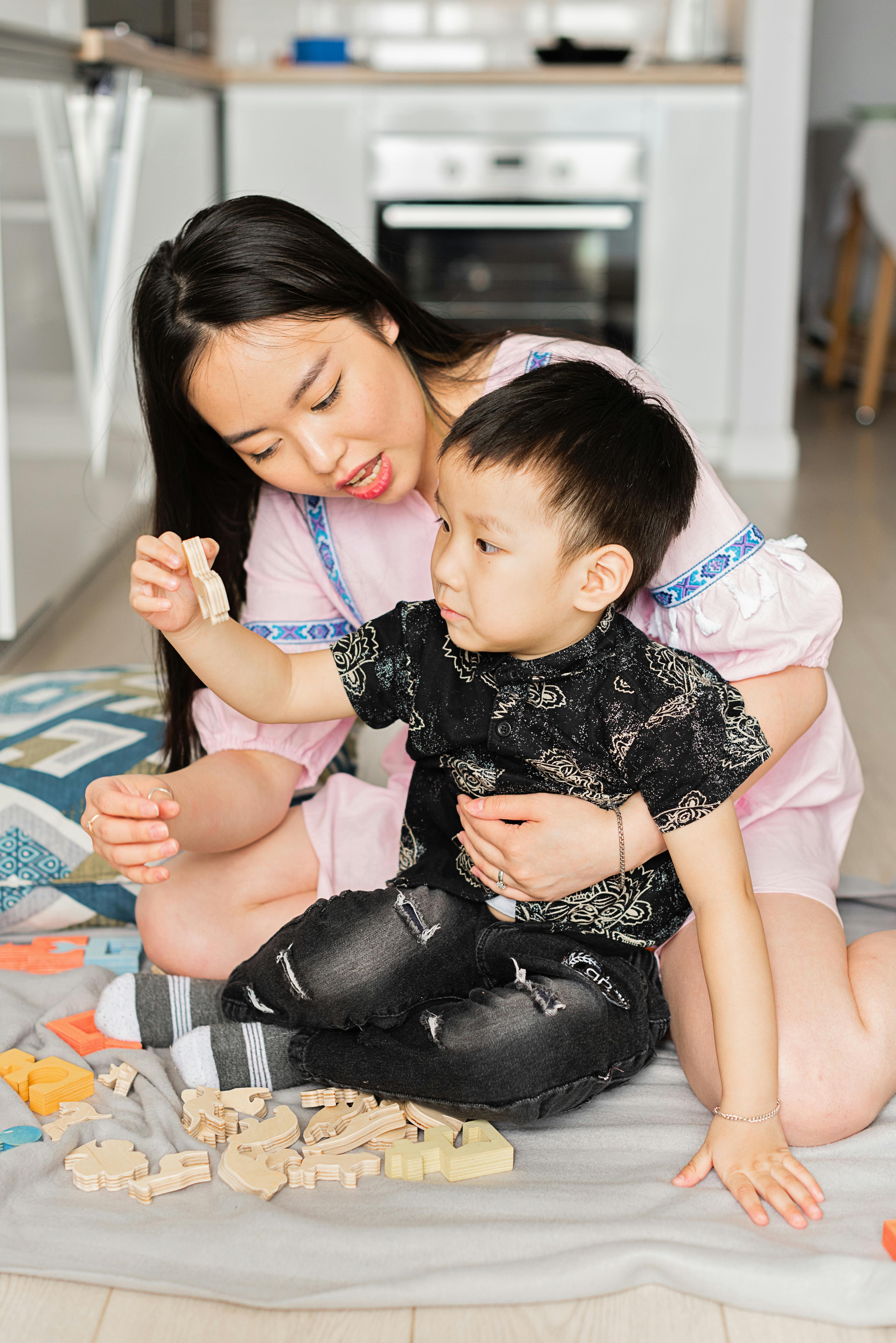 woman sitting behind boy in black clothes playing wooden toys on the floor