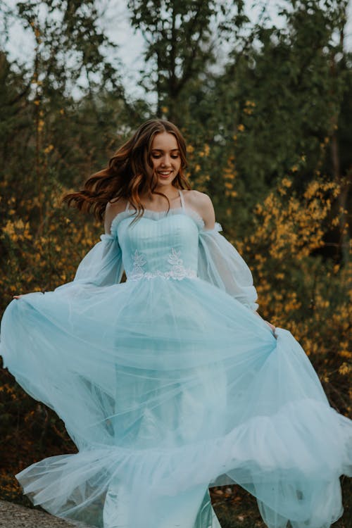 Cheerful female with flying wavy hair wearing elegant long light blue dress among plants in countryside