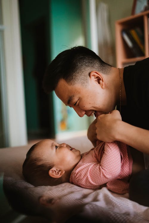Free Side view of cheerful Asian father hugging smiling little child lying in bed Stock Photo