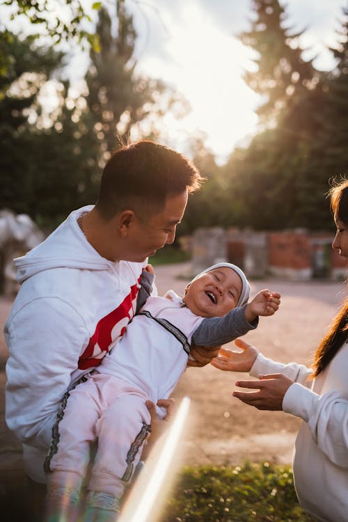 Free Cheerful young family holding little laughing baby in sunshine while walking in sunny garden Stock Photo