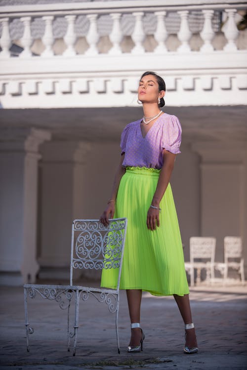 Woman in Purple Shirt and Green Skirt Standing beside White Metal Chair