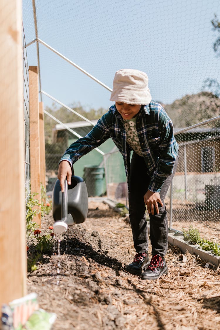 Boy In Plaid Shirt And Panama Hat Watering Soil