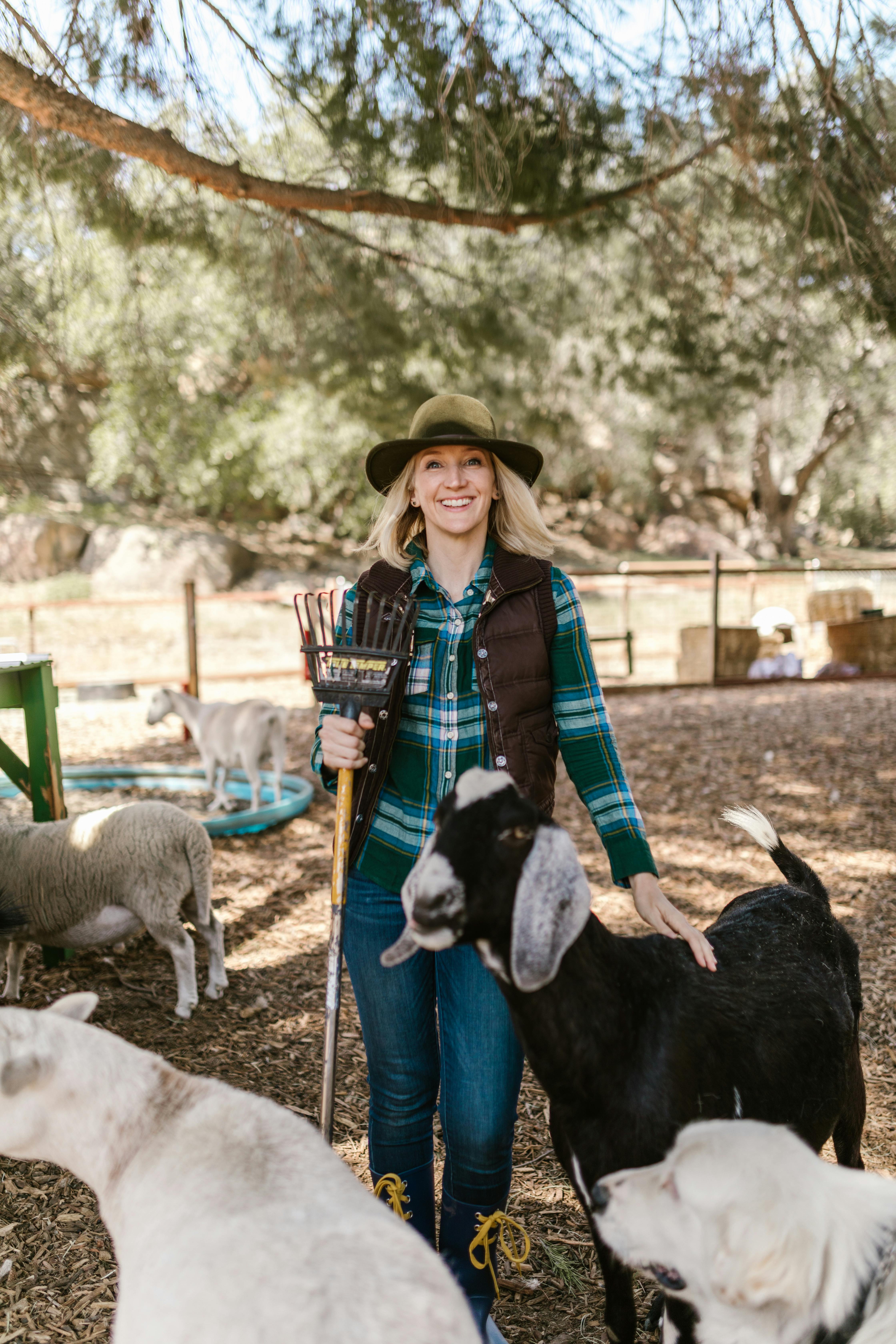 Cheerful woman in cowboy hat standing with goats on a sunny farm day.