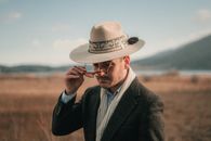 Confident male in elegant jacket and cowboy hat putting on eyeglasses in blurred countryside