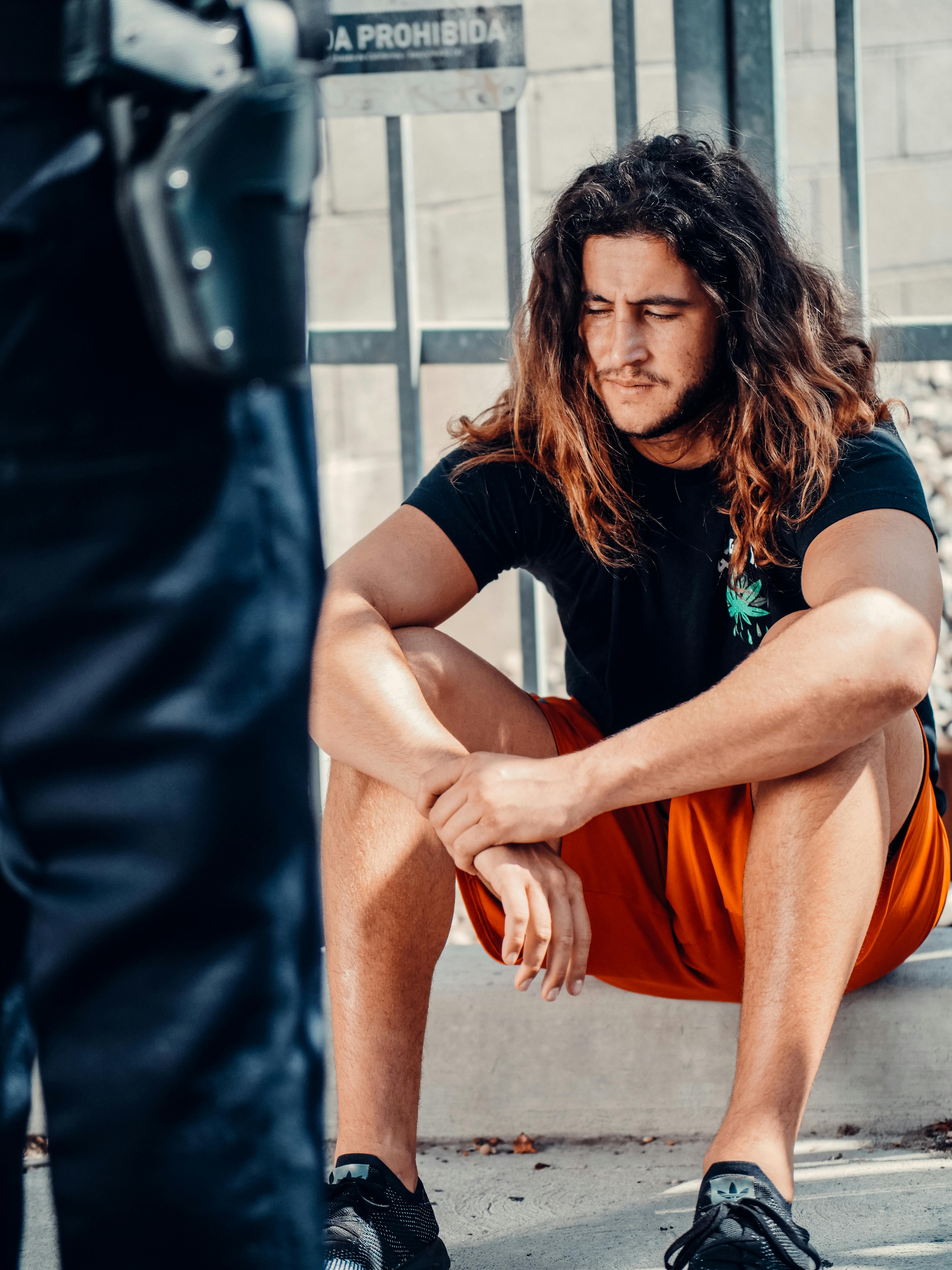 a man with long hair sitting on a pavement while looking down