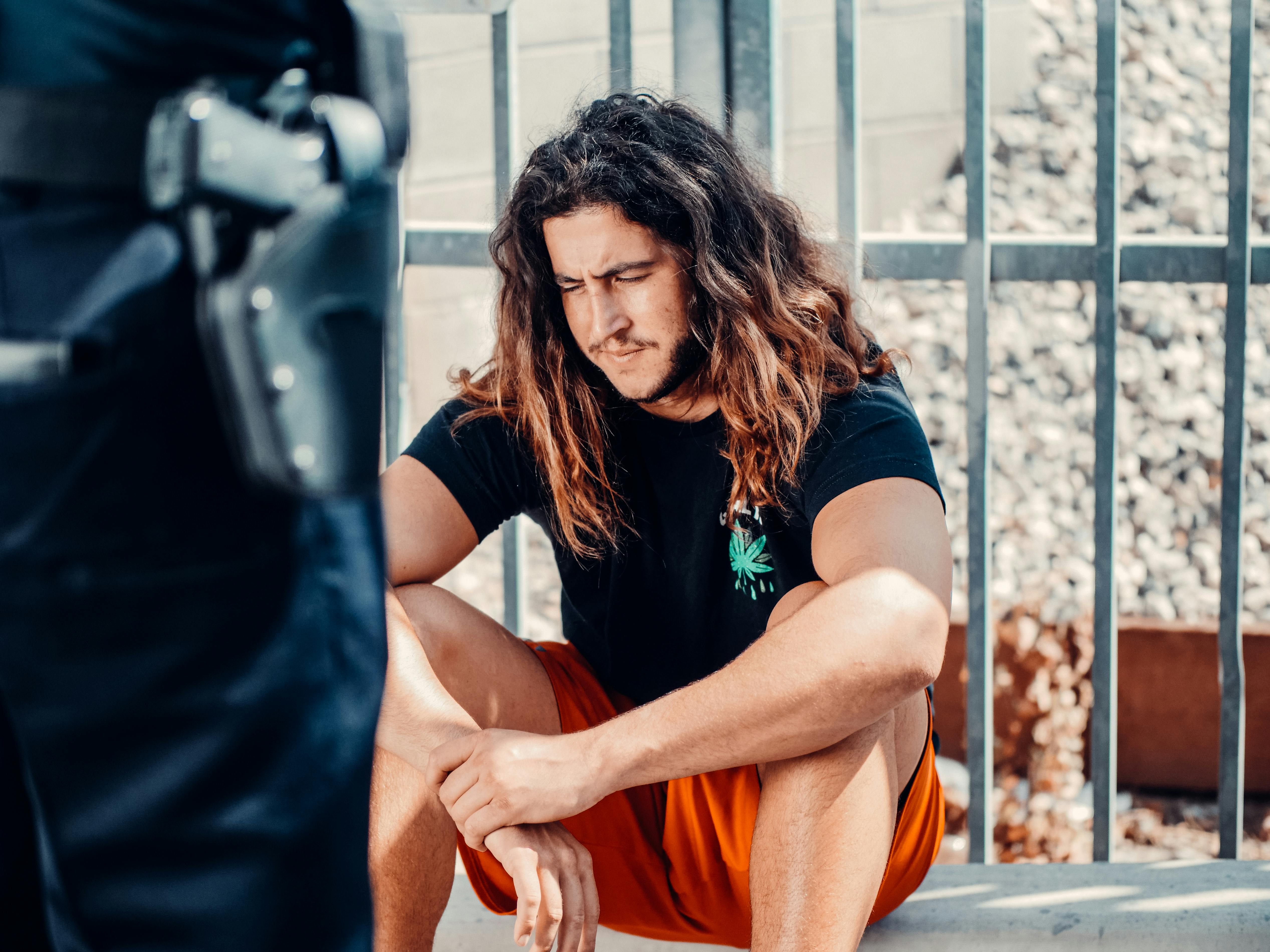 Man in black shirt with wavy hair sitting outdoors by a metal fence, looking contemplative.