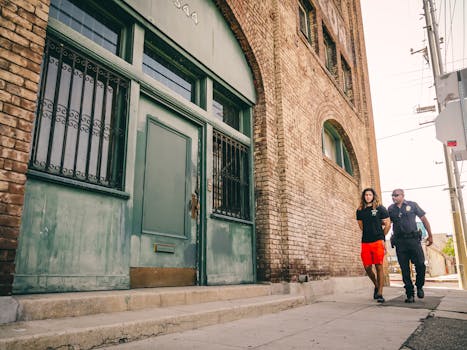 A police officer escorts a handcuffed individual past a brick building.