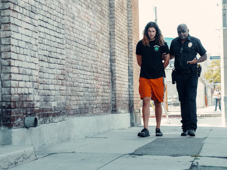 A Policeman And A Man With Long Hair Walking On The Street