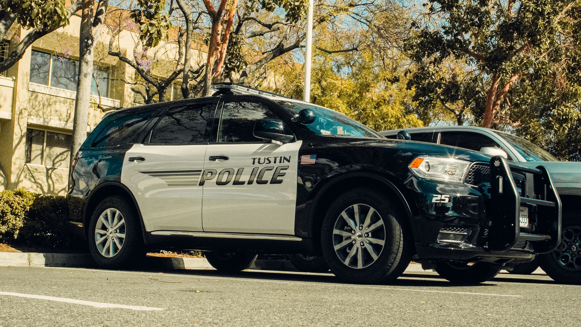 Tustin police car parked in a shaded lot surrounded by trees.