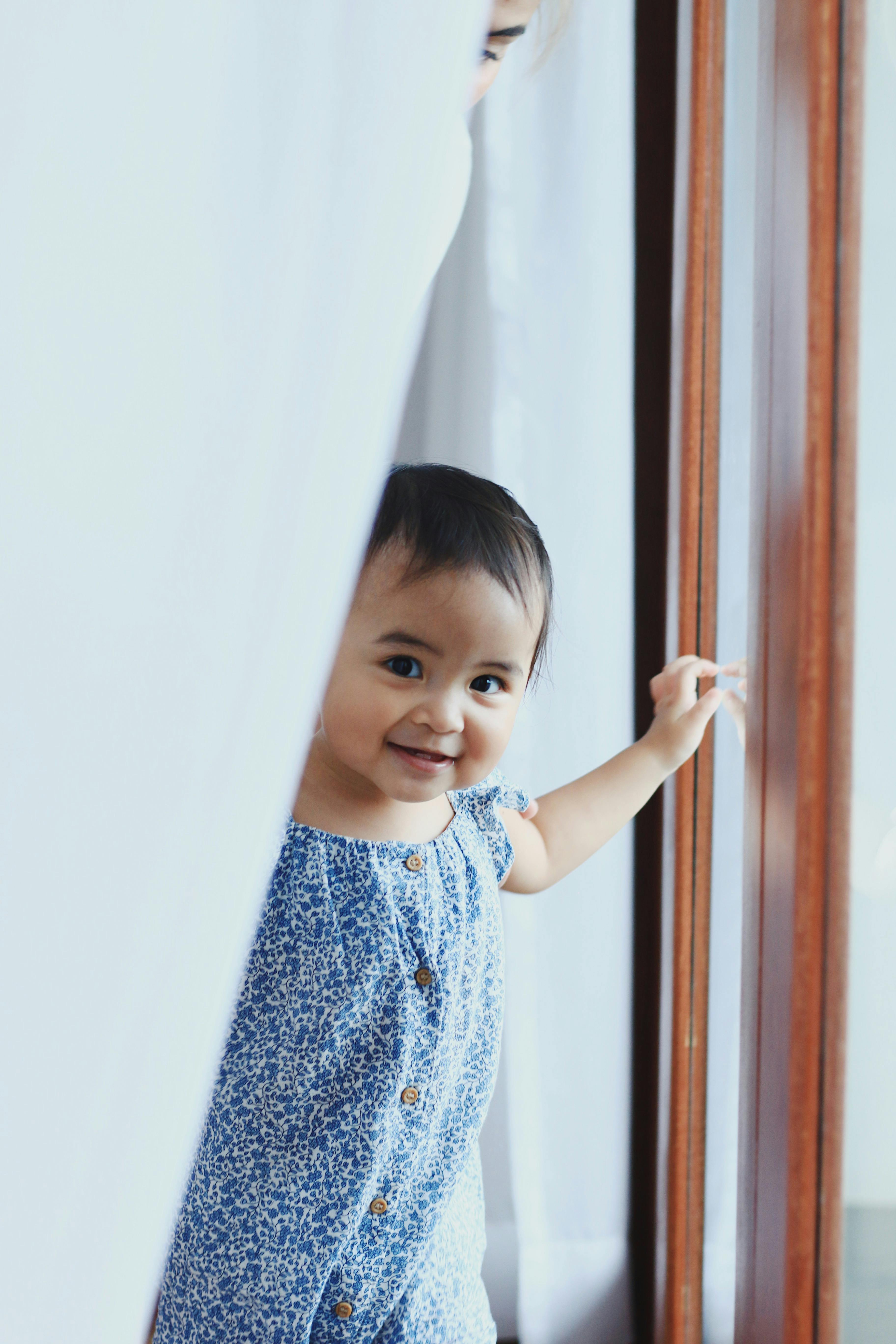 Adorable toddler in a blue dress smiles while peeking out from behind a curtain near a window.