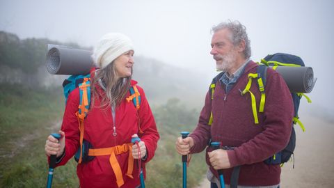 A senior couple hiking with trekking poles amidst foggy mountain landscape in Portugal, sharing a joyful moment.
