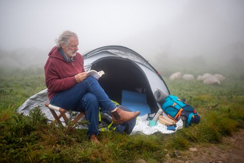 Free A senior man enjoys reading at a campsite in Portugal's lush landscape, surrounded by mist. Stock Photo