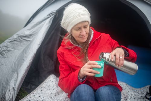 Free Caucasian woman in a red jacket camping, pouring a drink from a thermos by her tent in Portugal. Stock Photo