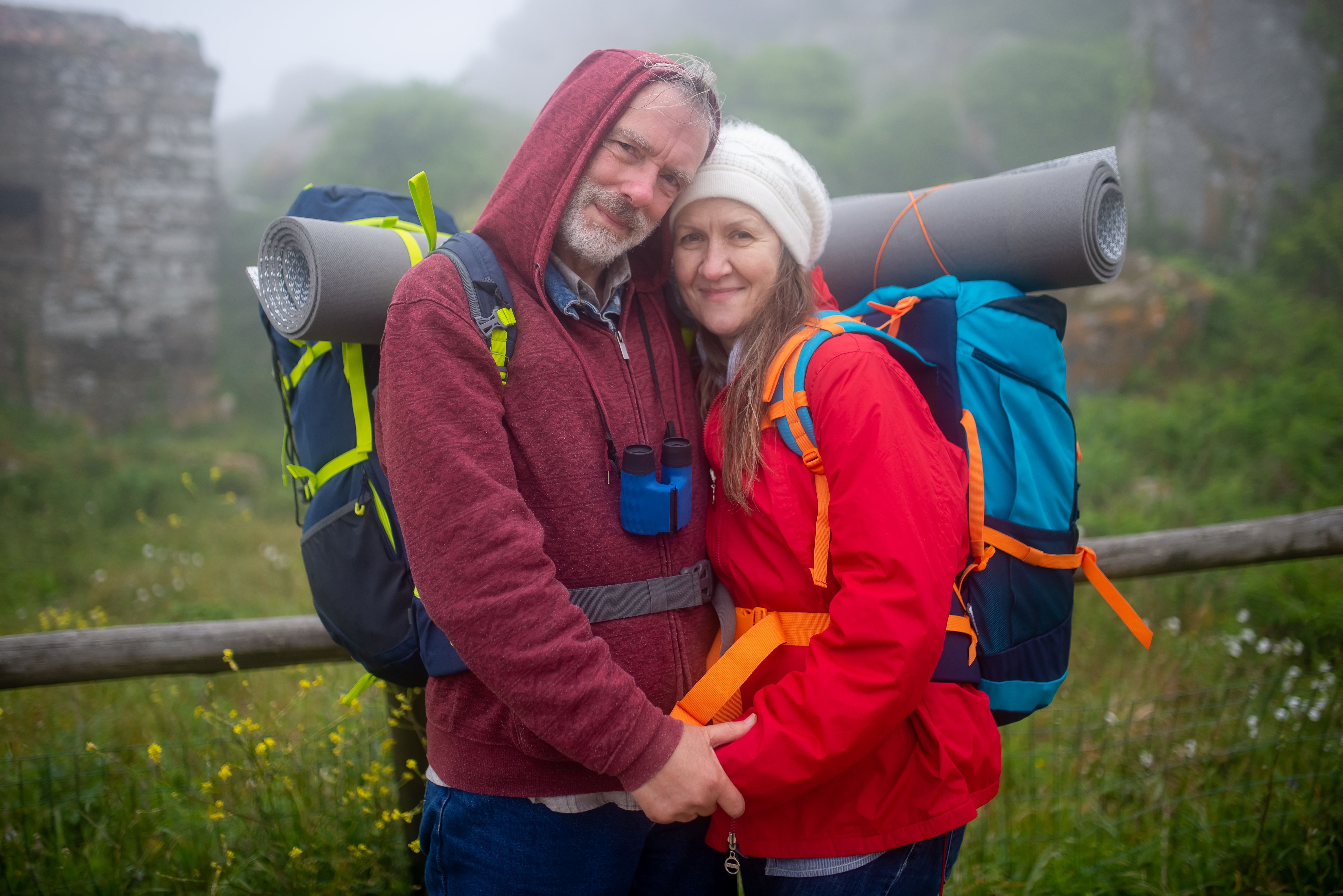 Elderly couple hiking in Portugal embraces with backpacks in a foggy landscape, showcasing love and adventure.