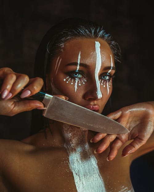 Woman model with glow bronzer and artistic makeup and wet hair covering mouth with kitchen knife