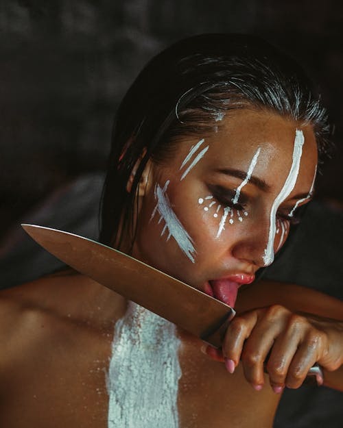 Model with artistic makeup touching knife with tongue