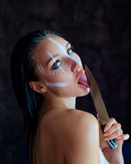 Side view of young female model with long dark wet hair bare shoulders with creative makeup with white stripes painted on face licking knife blade by tongue and mouth opened