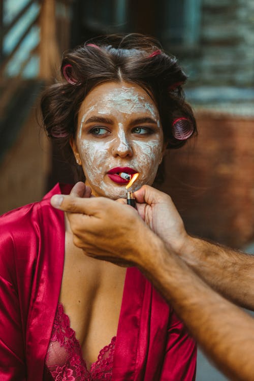 Young female with cosmetic face mask and hair curlers wearing lace underwear and satin dressing gown lighting up cigarette by lighter in male hands
