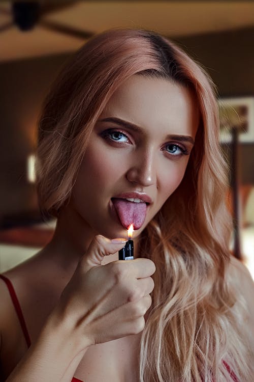 Young sensual female with light pink long hair touching lit lighter to tongue with open mouth