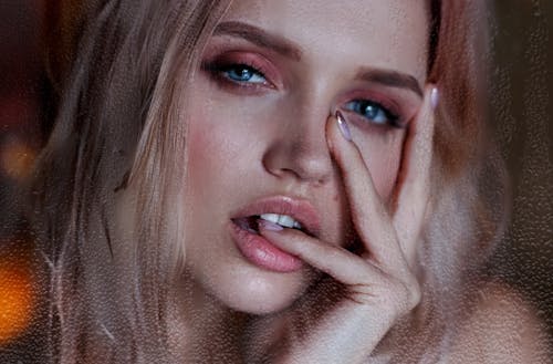Attractive young female with blue eyes pink eyeshadows and sensual lips biting finger and touching face with fingers and looking at camera
