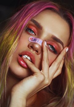 Close-up of a woman with pink hair and vibrant makeup, featuring 'love' text on a nose sticker.