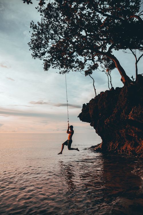 Free A person swings off a coastal cliff into the ocean against a sunset backdrop, creating a dramatic silhouette. Stock Photo