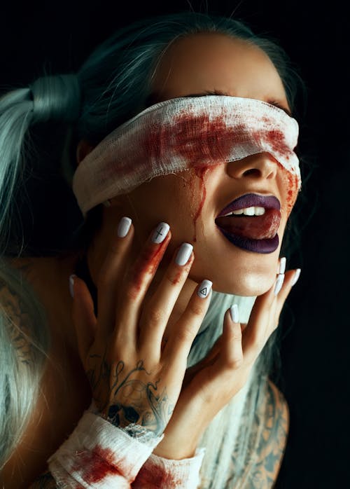 Young female with dyed hair and bloody gauze blindfold licking lips against black background
