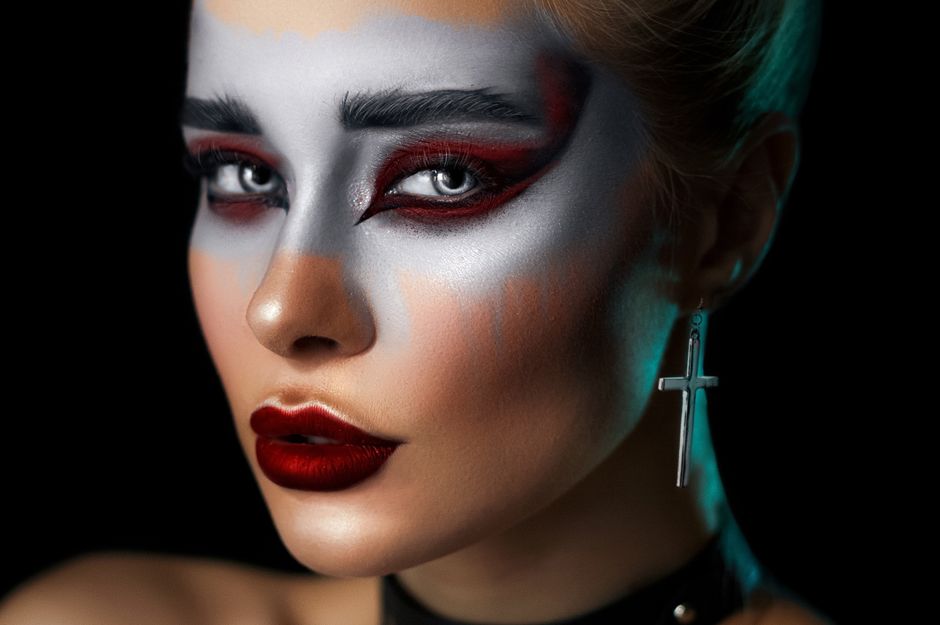 Young female model with creative makeup and stylish cross shaped earring looking at camera against black background