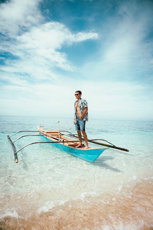 Barefoot male in shirt and shorts standing on bright boat floating on sea waves near wet shore against cloudy blue sky on resort
