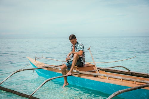 Barefoot man in shorts and shirt holding sunglasses and sitting on boat in clean azure sea on weekend day