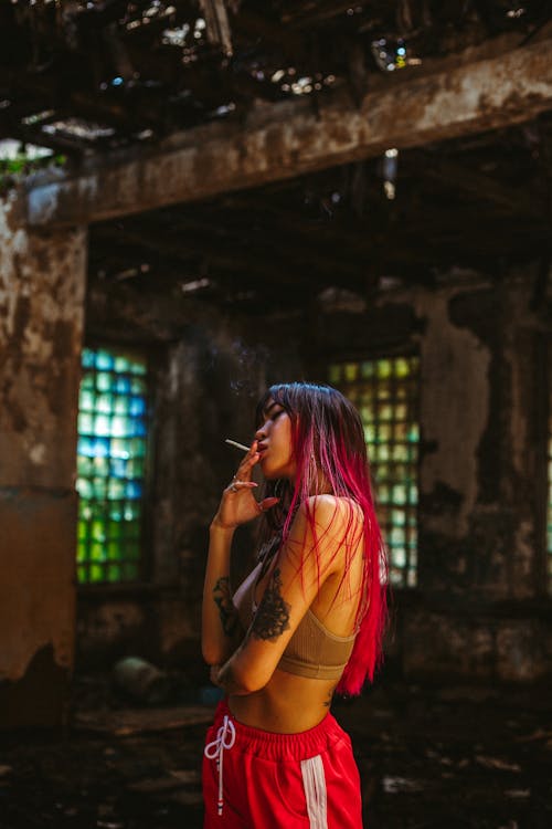 Free Side view young Asian woman with long pink hair and tattooed arms wearing topic and red sport pants smoking in destroyed building Stock Photo