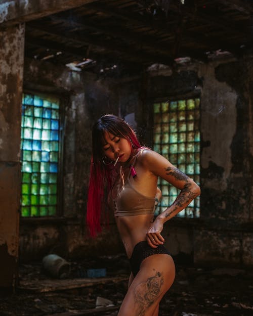 Side view of young sensual Asian woman in lingerie with tattoos on leg and arm and cigarette in mouth standing in shabby littered hangar