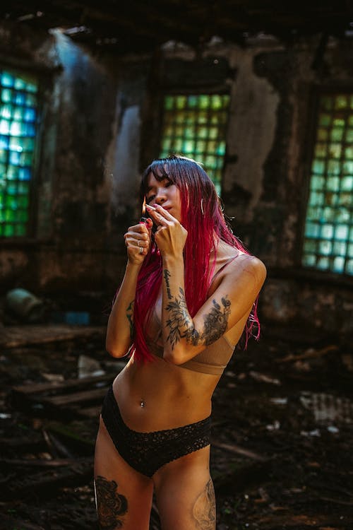 Tattooed Asian woman with long pink hair wearing lingerie lighting up cigarette in shabby hangar