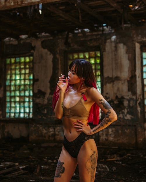 Concentrated young Asian female with long pink hair wearing black lace panties and top with tattooed arms and hips standing and smoking in abandoned hangar