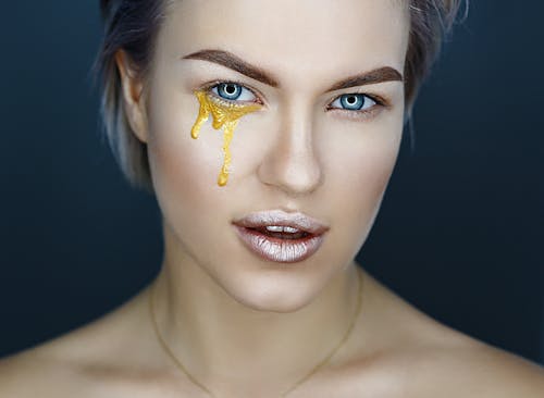 Female model with gold paint of artistic makeup under eye and metal lipstick on lips looking at camera