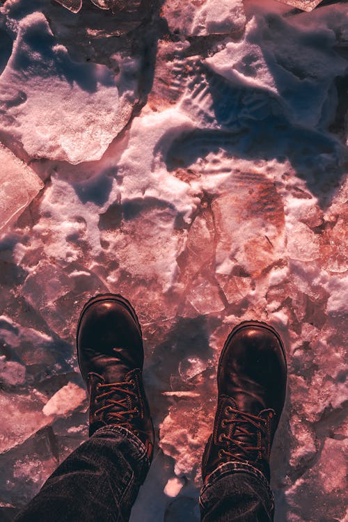 Free From above legs of person in warm leather boots and black jeans standing on frozen ground with dry leaves and snow in sunlight Stock Photo
