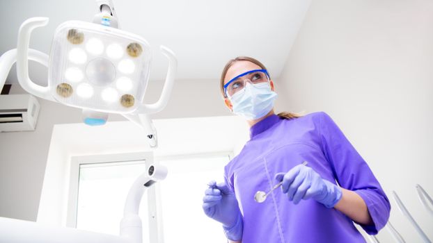 From below view of female dentist in medical uniform wearing mask and protective eyeglasses and latex gloves with dental mirror while preparing for examination of patient