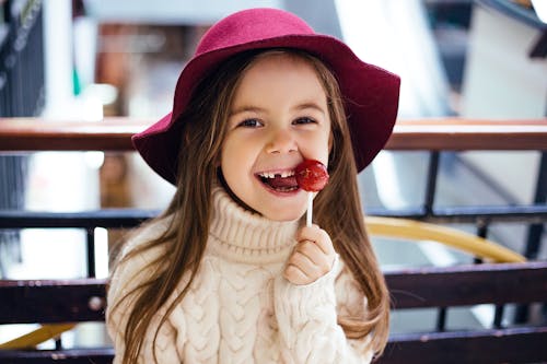 From above of cute little girl in sweater and hat standing with lollipop near railing while smiling and looking at camera