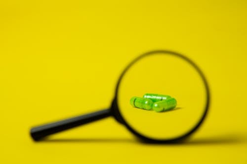 Closeup of magnifying glass placed near heap of green pills on bright yellow surface in studio