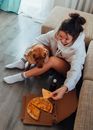 From above of young female owner hugging obedient purebred Labrador while sitting on floor near sofa and eating pizza during weekend at home