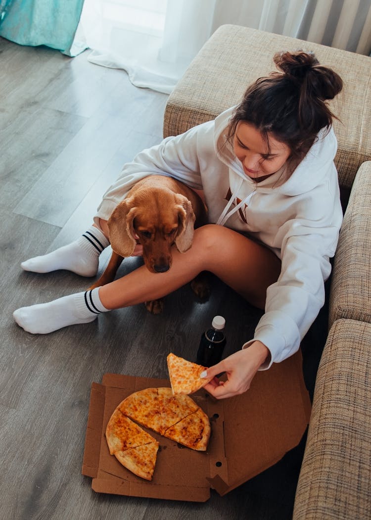 Woman Sitting With Dog On Floor And Eating Pizza