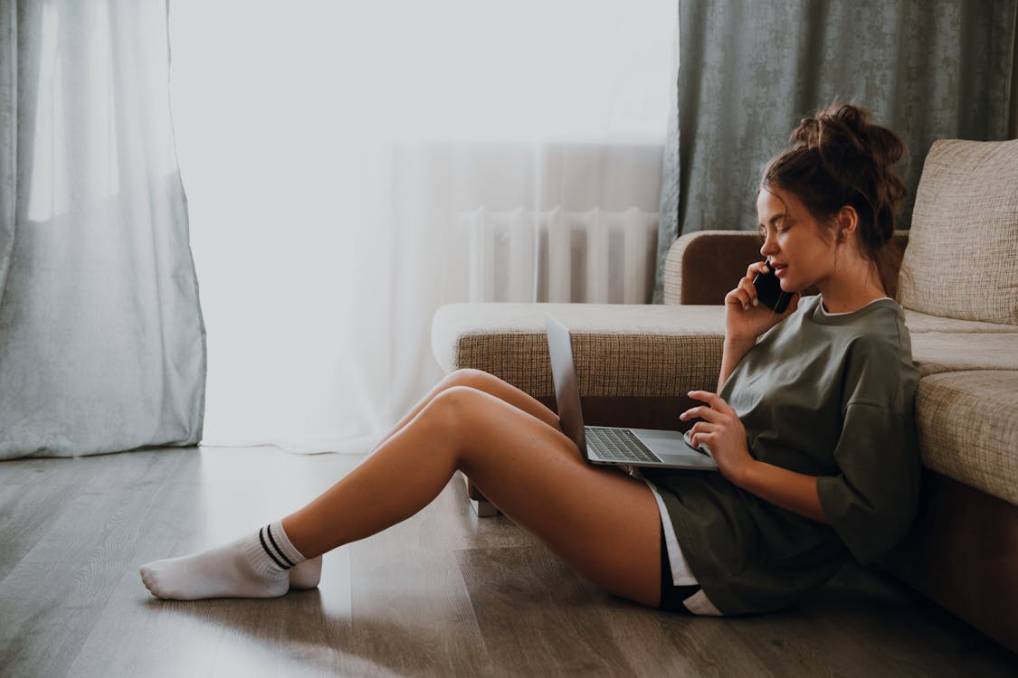 Side view of young female in domestic outfit sitting on floor near sofa and browsing netbook while talking on mobile phone at home