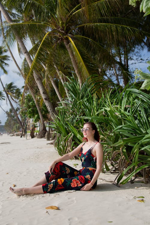 Side view of smiling young barefoot female with long brown hair and in bright bodysuit sitting on sandy shore near tropical palms and looking away during vacation